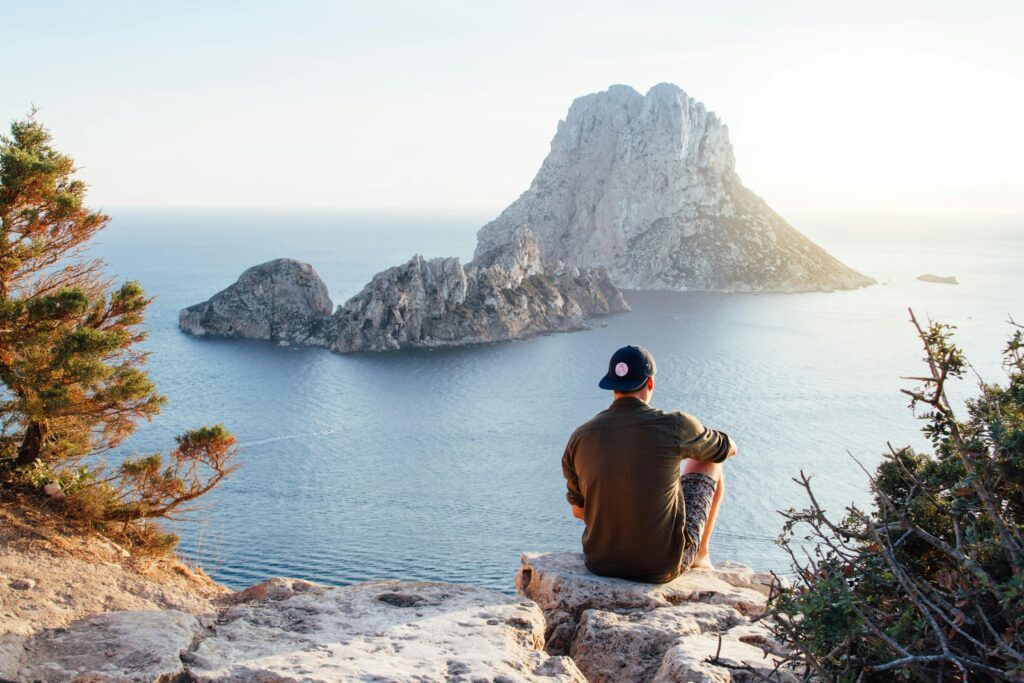 A man sitting on the cliff overlooking the ocean with big rocky island in the background.