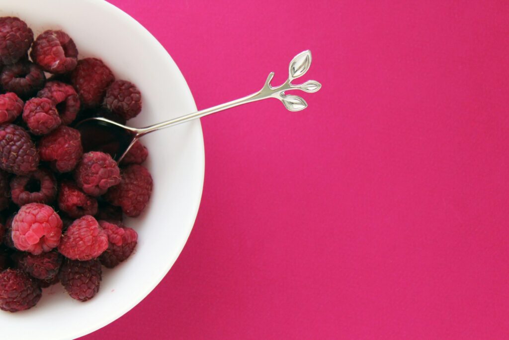 A bowl of raspberries with a silver spoon on a fuchsia background